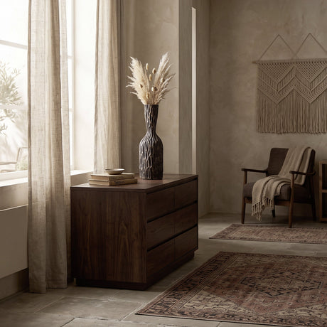 Sakti Vase in black and gold, tall bottle shape with faceted ridges, displayed on a dark wood dresser with dried pampas grass in a warm living room setting.