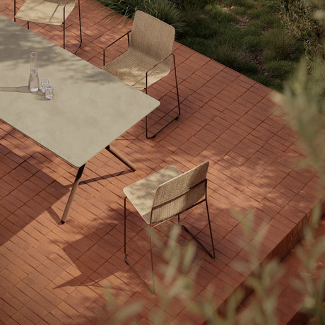 Sal Steel Stacking Garden Dining Armchair with woven beige seat and back on a slim steel sled frame, shown on a terracotta tiled outdoor patio beside a dining table.