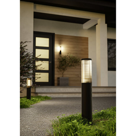 Salle Bollard Light in black aluminium and plastic, shown lit on a garden path beside a modern home entrance, with its ribbed smoked shade glowing above a slim cylindrical base.
