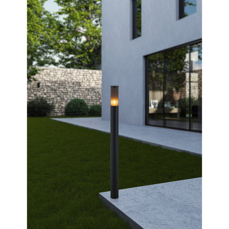 Salle Bollard Light in black aluminium with a ribbed smoked shade, illuminated on a garden lawn beside a paved patio and modern house.