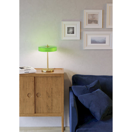 Salve Table Lamp in brushed brass with a glossy green opal glass disc shade, shown lit on a wooden sideboard beside a navy sofa and framed wall art.