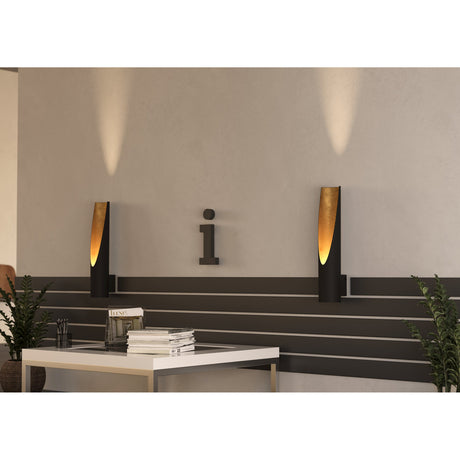 San Cono Wall Light 11cm in black and gold, slim steel cylinder with angled cut revealing a glowing gold interior, mounted on a wall above a living room coffee table.