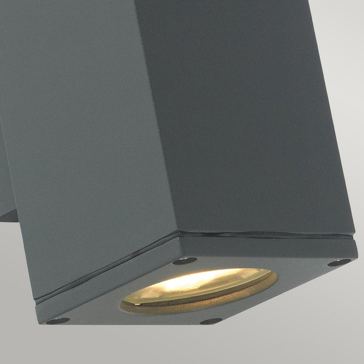 A close-up view of the Sandvik Outdoor 2 Light Up & Down Wall Light in graphite, featuring a black angular design against a gray background. This IP65-rated fixture is angled downward to cast a warm glow, showcasing its sleek, modern hexagonal shape.
