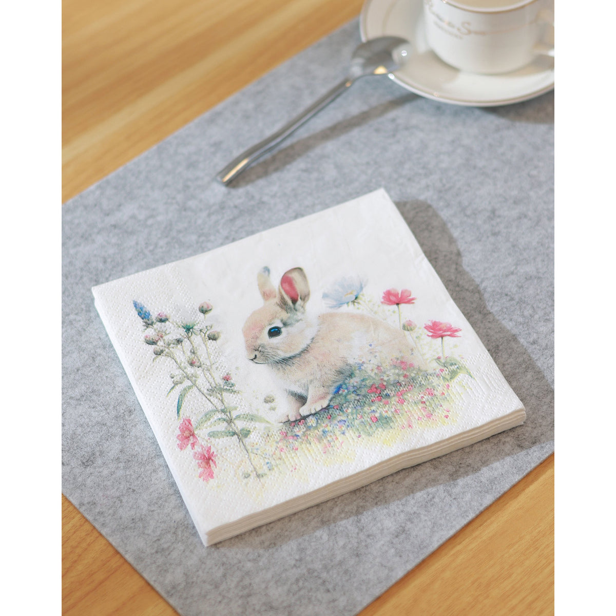 Sasaram Napkins Set of 20, white paper napkin printed with a rabbit among wildflowers, folded square on a grey mat beside a teacup and spoon on a wooden table.