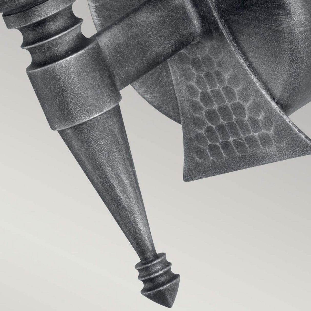 Close-up of the Saxon 1 Light Wall Light, highlighting its metal structure with textured, geometric, and pointed features. It showcases a hexagonal pattern on its flat surface. The fixture's angled, tapered rod with ornate details is finished in a silver patina against a light, neutral background.