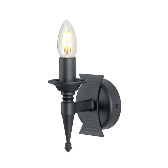 The Saxon 1 Light Wall Light - Black features a black wall sconce with a single faux candle-shaped bulb, complemented by a round backplate and textured, angled support. Its wrought iron frame and medieval-inspired design provide a traditional yet elegant look.