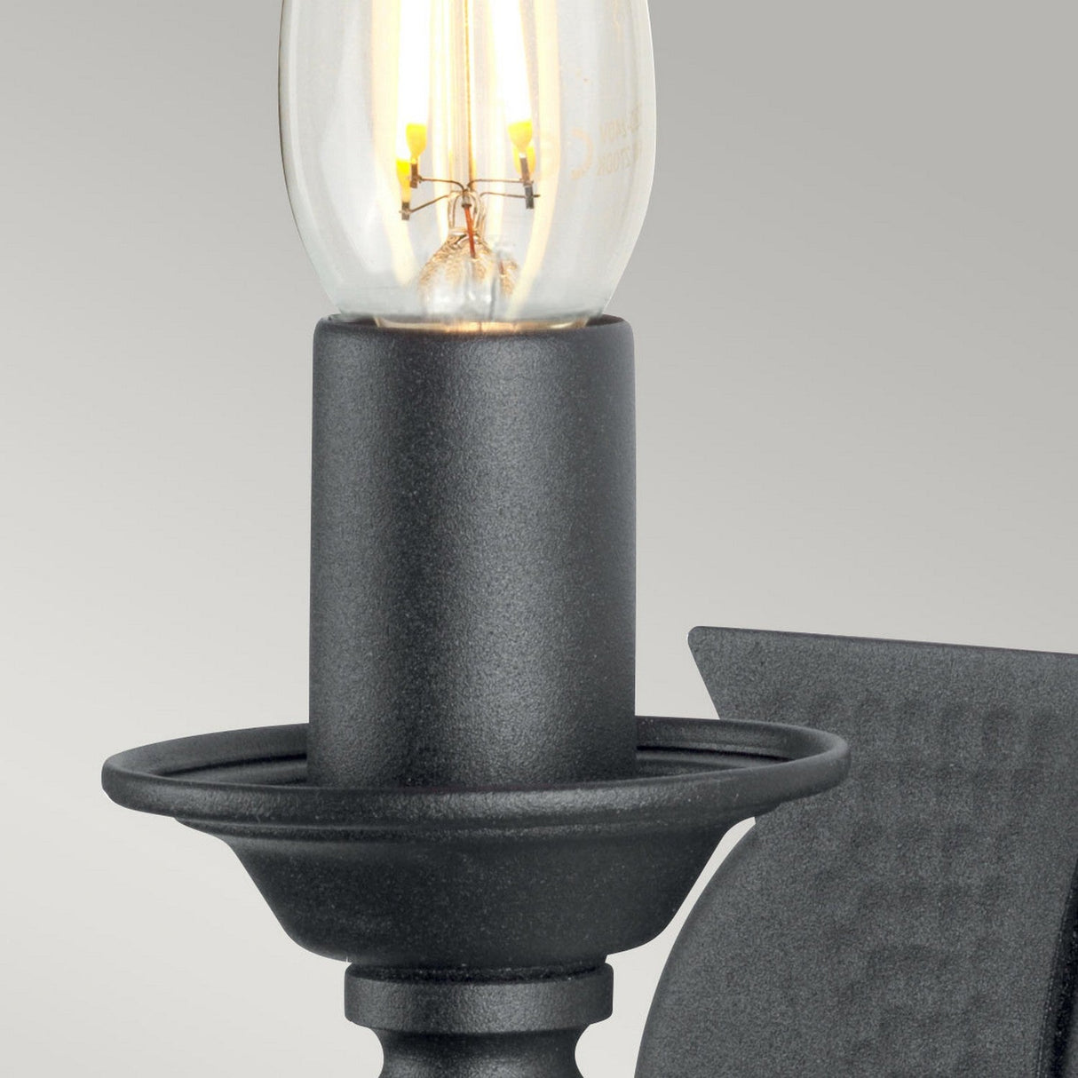 Close-up of the Saxon 1 Light Wall Light - Black featuring a wrought iron frame and an exposed bulb. The simple, light gray background highlights its elegant curves and contemporary finish, echoing a subtle medieval-inspired design.