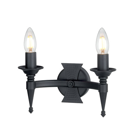 The Saxon 2 Light Wall Light - Black is a wall-mounted wrought iron fixture with two candle-style bulbs flanking a central circular base. Its design incorporates pointed elements and a textured symmetrical cross pattern, reminiscent of medieval-inspired lighting aesthetics.