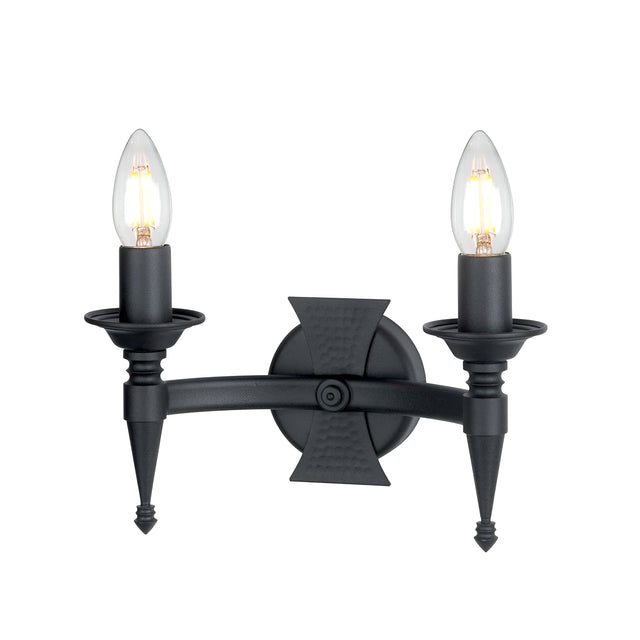 The Saxon 2 Light Wall Light - Black is a wall-mounted wrought iron fixture with two candle-style bulbs flanking a central circular base. Its design incorporates pointed elements and a textured symmetrical cross pattern, reminiscent of medieval-inspired lighting aesthetics.