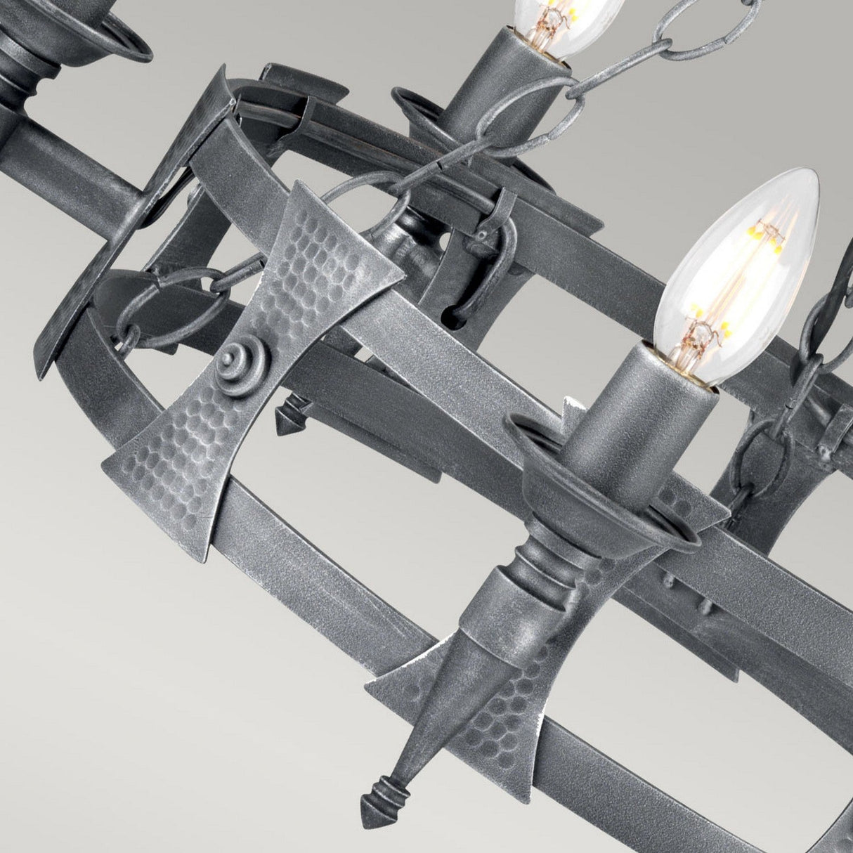 The Saxon 5 Light Chandelier in Black/Silver Patina showcases a medieval-style design with its close-up view highlighting the intricate wrought iron elements that resemble crossed arrows. This chandelier holds five candle-shaped bulbs, casting a warm glow and enhancing its timeless elegance.