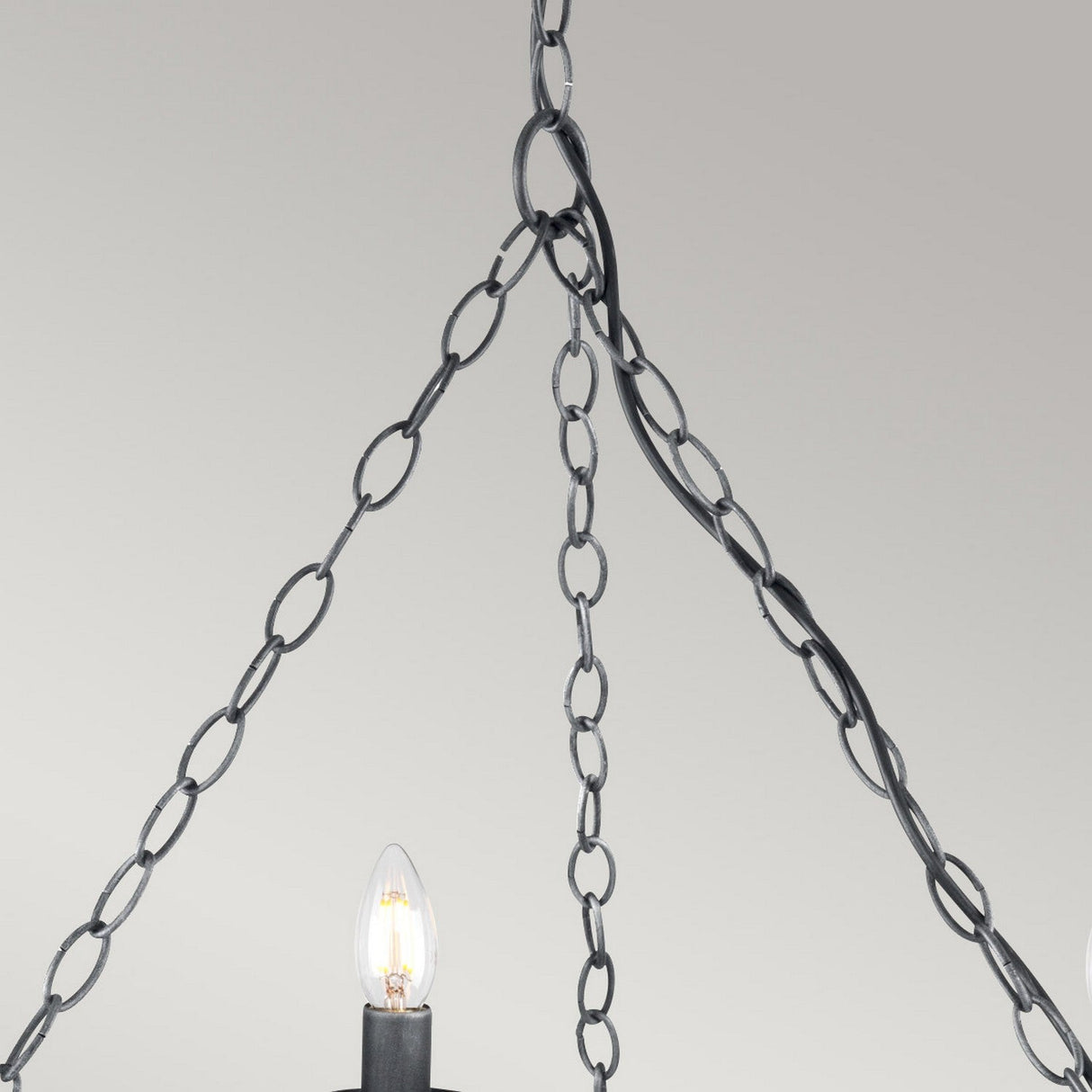 Close-up of the Saxon 8 Light Chandelier - Black/Silver Patina, showcasing its medieval-inspired craftsmanship with three black metal chains converging at the top. Two candle-like bulbs on black stands emit a soft glow against a light gray background, highlighting its wrought iron elegance.