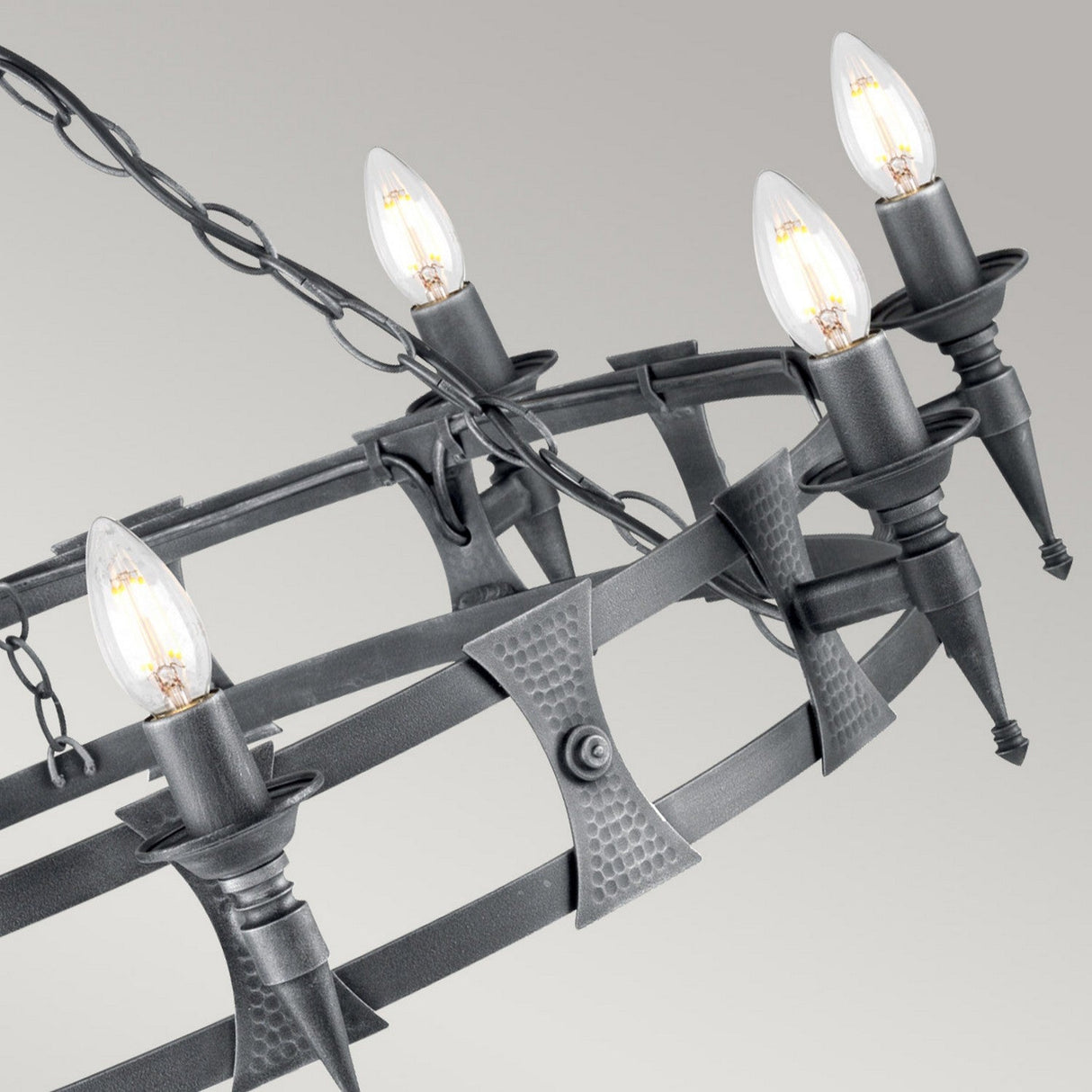 A close-up of the Saxon 8 Light Chandelier - Black/Silver Patina highlights its rustic, industrial aesthetic with eight candle-shaped light bulbs arranged on a wrought iron frame. The chandelier's chain and angular metal design bring to mind medieval craftsmanship, providing both a contemporary and vintage feel against a grey background.