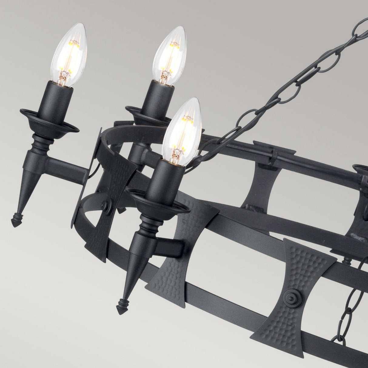 The Saxon 8 Light Chandelier - Black features a circular frame and hangs on a chain, blending medieval design elements with modern aesthetics. Crafted from wrought iron, it showcases eight decorative arms that support candle-style bulbs, creating a rustic yet contemporary look against neutral settings.