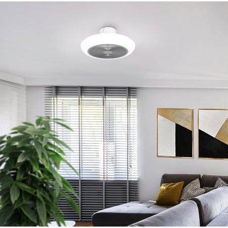 Sayulita ceiling fan with light in white and grey, round low-profile design with acrylic shade, mounted in a living room above a sofa and window blinds.