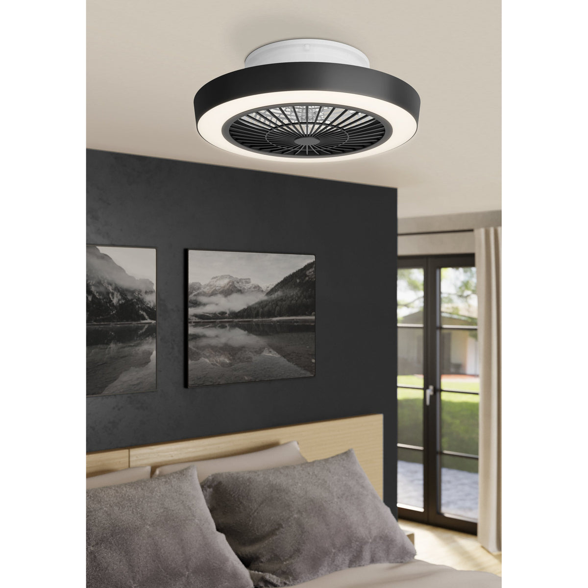 Sazan ceiling fan with light in black and white, round flush-mount fitting with black outer band, acrylic diffuser ring and black radial grille, shown above a bed in a dark-walled bedroom.