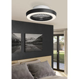 Sazan ceiling fan with light in black and white, round flush-mount fitting with black outer band, acrylic diffuser ring and black radial grille, shown above a bed in a dark-walled bedroom.
