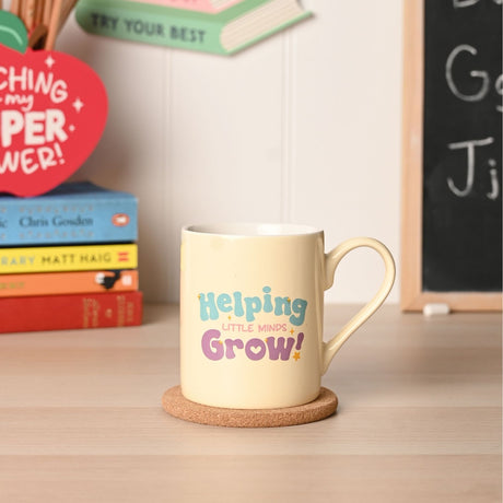 Cream porcelain mug with curved handle printed with "Helping Little Minds Grow!" in teal, pink and purple, shown on a cork coaster beside stacked books on a desk.