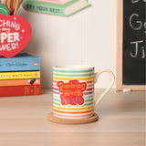 Porcelain mug with a teal, green, yellow, orange, pink and purple rainbow stripe, cream body and red heart badge reading Teaching Is A Work Of Heart, on a cork coaster.