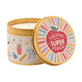 Round gold tin scented candle with lid, cream label printed with classroom items, lid featuring a red apple and "Teaching is my super power" text on pastel stripes.