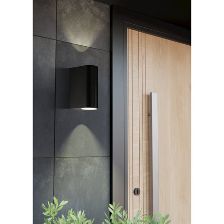 Scogliera-E Outdoor Wall Light in black plastic, mounted beside a wooden front door, casting light upwards and downwards from its compact rounded-rectangle body.