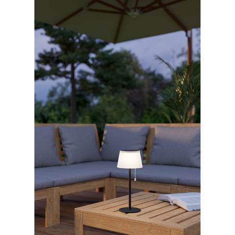 Scordiano Solar LED Table Lamp in black with a white pleated shade on a wooden coffee table, set against an outdoor lounge area with grey cushioned seating.