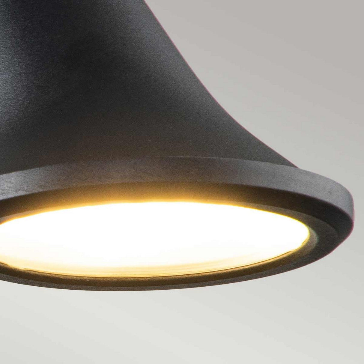 A close-up of the Selma Outdoor LED Wall Light in black, turned on and casting a warm glow. The backdrop is a plain, neutral grey that elegantly highlights the illuminated underside. Echoing the precision of architectural lighting design, it serves as an exquisite focal point in any space.