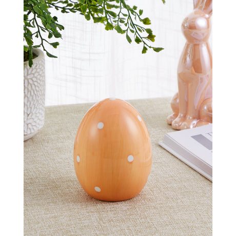 Sendayan ceramic decorative egg in glossy pastel apricot with white polka dots, displayed upright on a neutral table beside a plant and pink rabbit figurine.