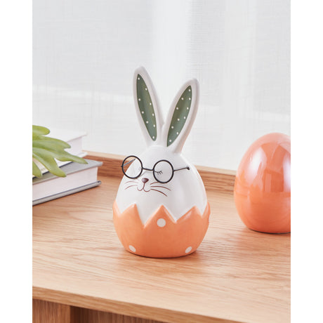 White ceramic rabbit ornament with egg-shaped body, cracked orange eggshell base with white polka dots, green polka-dot ears, and round black glasses, displayed on a wooden shelf near books.