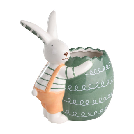 Ceramic white rabbit ornament in a green-striped top and orange dungarees hugging a sage-green egg-shaped pot with a zigzag rim and white loop patterns, on a plain white background.