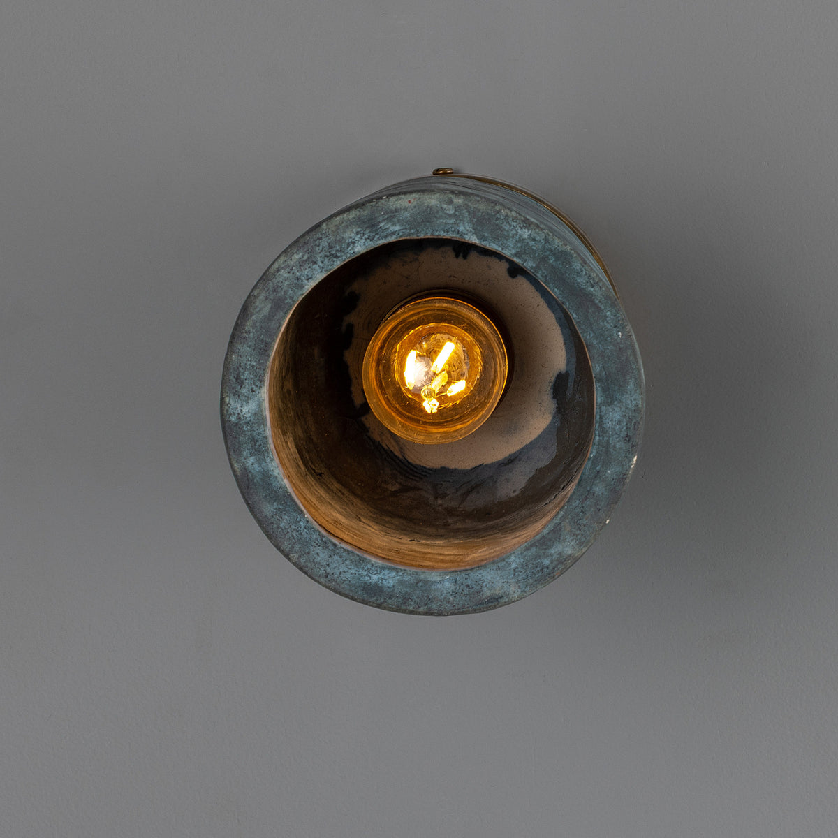 Senna Ceramic Ceiling Light in Blue Earth glaze, round cylinder shade with dark mottled blue-green finish, brass canopy, glowing E27 bulb visible inside, shown against a plain grey background.