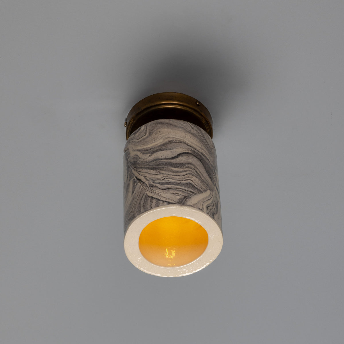 Senna Ceramic Ceiling Light with grey and cream marbled cylindrical shade, brass ceiling canopy, and glowing open base, shown against a plain grey background.