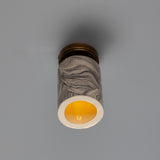Senna Ceramic Ceiling Light with grey and cream marbled cylindrical shade, brass ceiling canopy, and glowing open base, shown against a plain grey background.