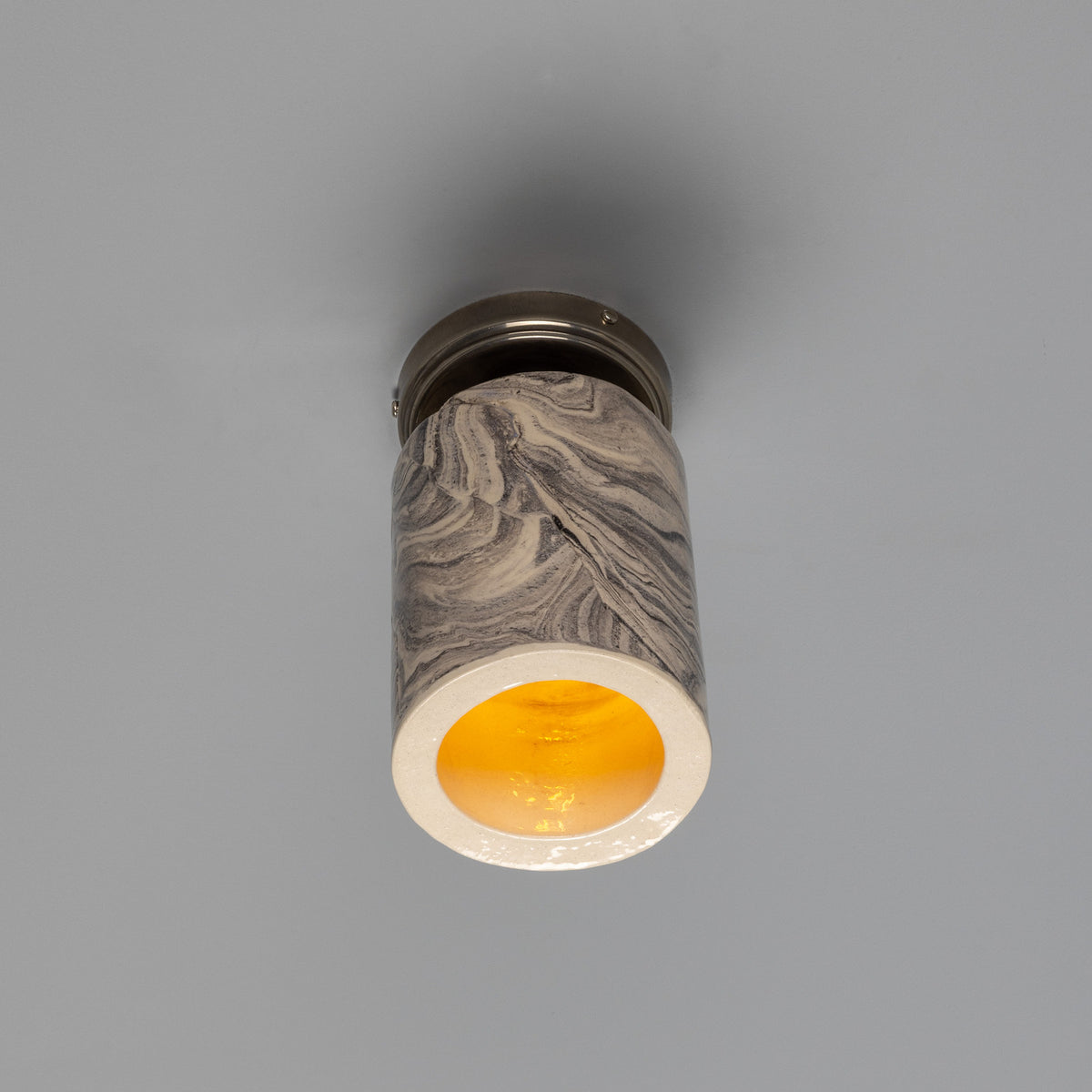 Senna Ceramic Ceiling Light in marbled grey and cream glaze, slim cylindrical shade with open base glowing amber, mounted on a round silver canopy against a plain grey background.