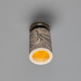 Senna Ceramic Ceiling Light in marbled grey and cream glaze, slim cylindrical shade with open base glowing amber, mounted on a round silver canopy against a plain grey background.