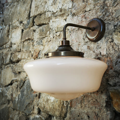 Seralba Glass Wall Light IP44 with a broad white schoolhouse-shaped shade in a brass gallery, curved brass arm and round wall plate, mounted on a stone wall.
