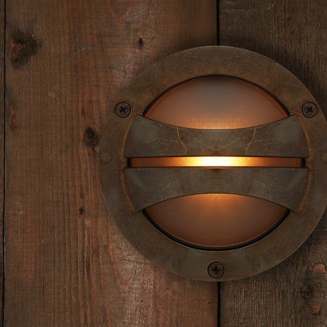 Seri Brass Outdoor Bulkhead Light IP54, round 14cm fixture with frosted glass and curved brass bar, mounted on a dark wood plank wall, lit to show a glowing centre slot.