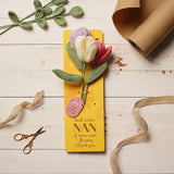 Crochet tulip flower bundle with lilac, cream, and pink blooms on sage-green stems, mounted on a yellow "Best Ever Nan" message card, styled with ribbon and kraft paper.