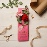 Crochet rose flower bundle with pink and red blooms on sage green stems, mounted on a pink "Happy Birthday" card, styled on a white wood surface with ribbon and kraft paper.