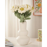 Crochet peony stems, 35cm, with white curled petals and yellow centres on green leafy stems, arranged in a white bulbous vase on a table.
