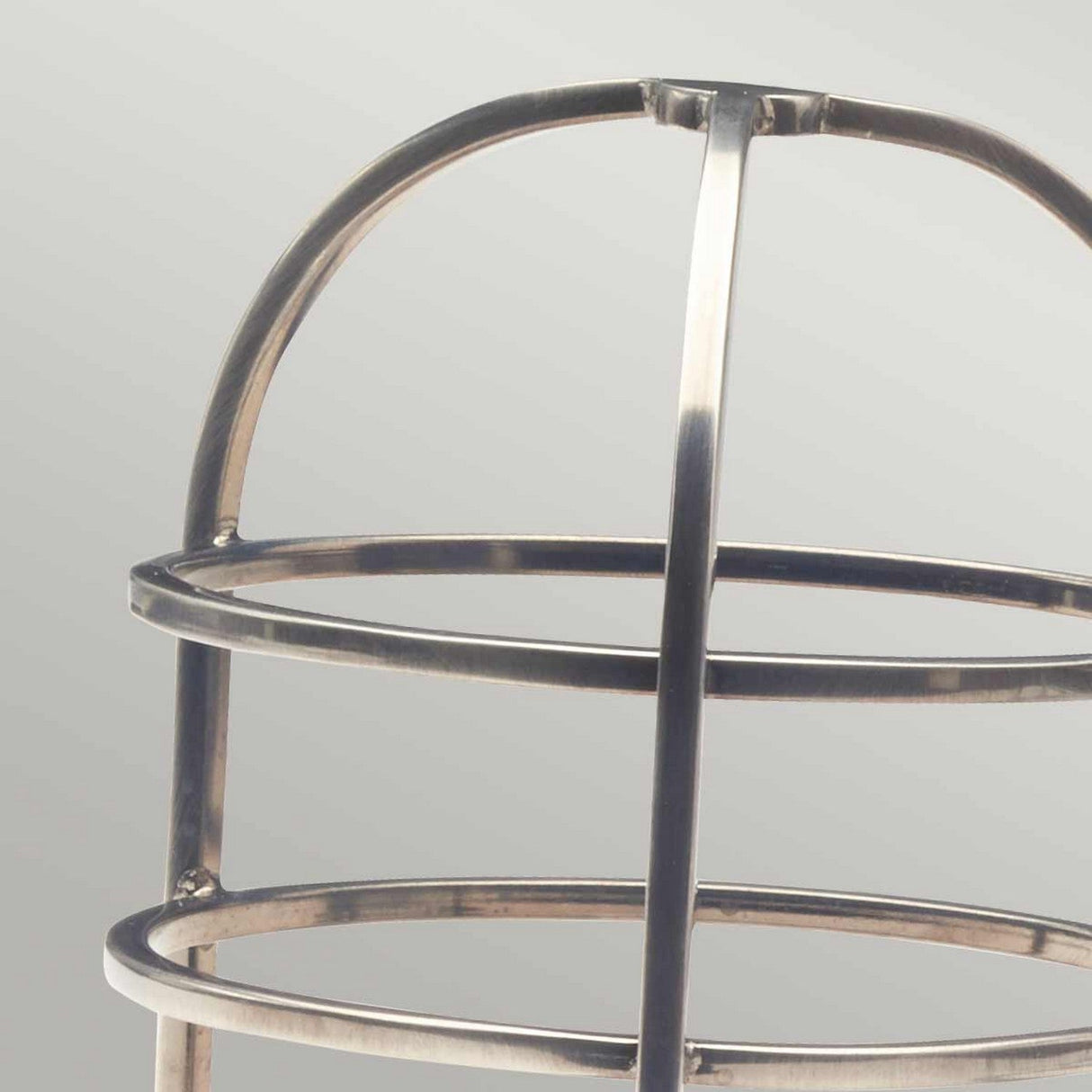 The close-up features the Cage Accessory For Sheldon & Somerton in Antique Nickel on a light gray background, highlighting its elegantly curved bars that form a dome shape at the top. This modern lighting design consists of two circular rings and vertical supports, reminiscent of traditional lantern protection.