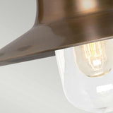A close-up of the Sheldon 1 Light Outdoor Wall Light in aged brass exudes industrial charm with its metal shade. Its clear glass enclosure highlights the glowing filament bulb, enhancing its vintage appeal against a plain, softly lit background.