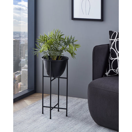 Shikabe metal plant stand in black with a round bowl on a slim four-legged tubular frame, shown holding a green potted plant in a grey living room with a sofa and window view.