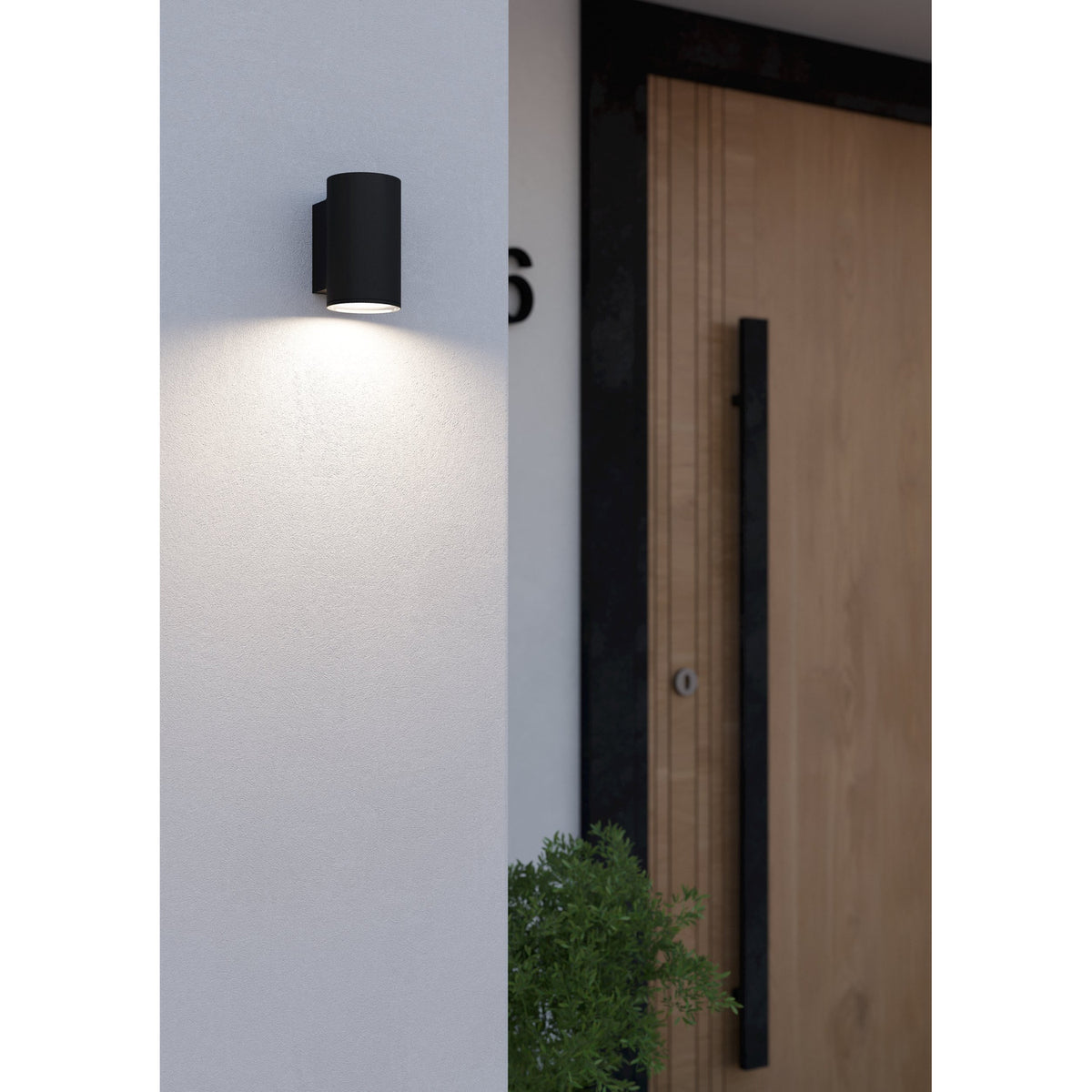 Silvessa Outdoor Wall Light in black steel, a cylindrical downlight mounted beside a wooden front door, casting warm downward light onto a light grey wall.