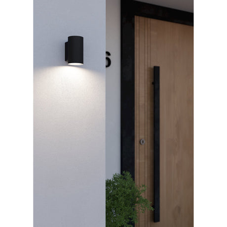 Silvessa Outdoor Wall Light in black steel, a cylindrical downlight mounted beside a wooden front door, casting warm downward light onto a light grey wall.
