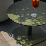 Sirkel Aluminium Garden Side Table in green, round top with rounded square base and central leg, shown outdoors with dappled shade and a drink glass on the surface.