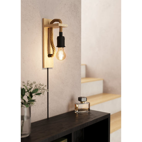 Sirvena wall light with wooden backplate and arm, twisted rope detail, and black E27 lampholder with exposed bulb, shown lit beside a staircase in a hallway setting.