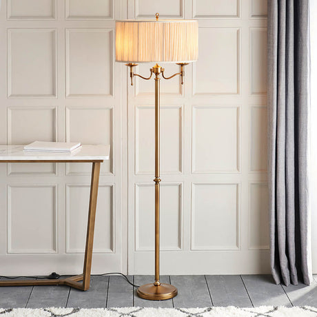 Interiors 1900 Ambledale antique brass floor lamp with twin E14 candle lights and champagne fabric shade, beside a marble table and grey curtain.