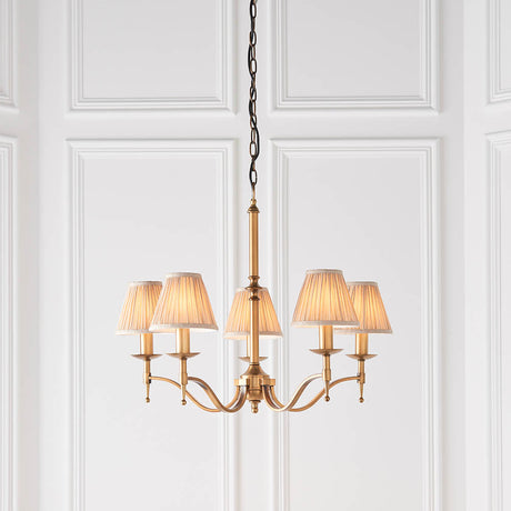 Interiors 1900 Ambledale 5 light chandelier in antique brass with curved arms and champagne fabric candle shades, shown against white panelled walls.