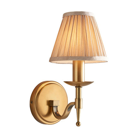 Interiors 1900 Ambledale single wall light in antique brass with a curved arm and champagne pleated fabric candle shade, on a white background.