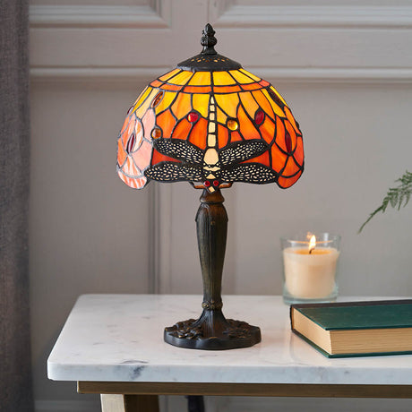 Alderworth Mini Tiffany table lamp with amber flame leaded glass shade and dragonfly motif on a dark base, set on a marble table beside a candle and book.